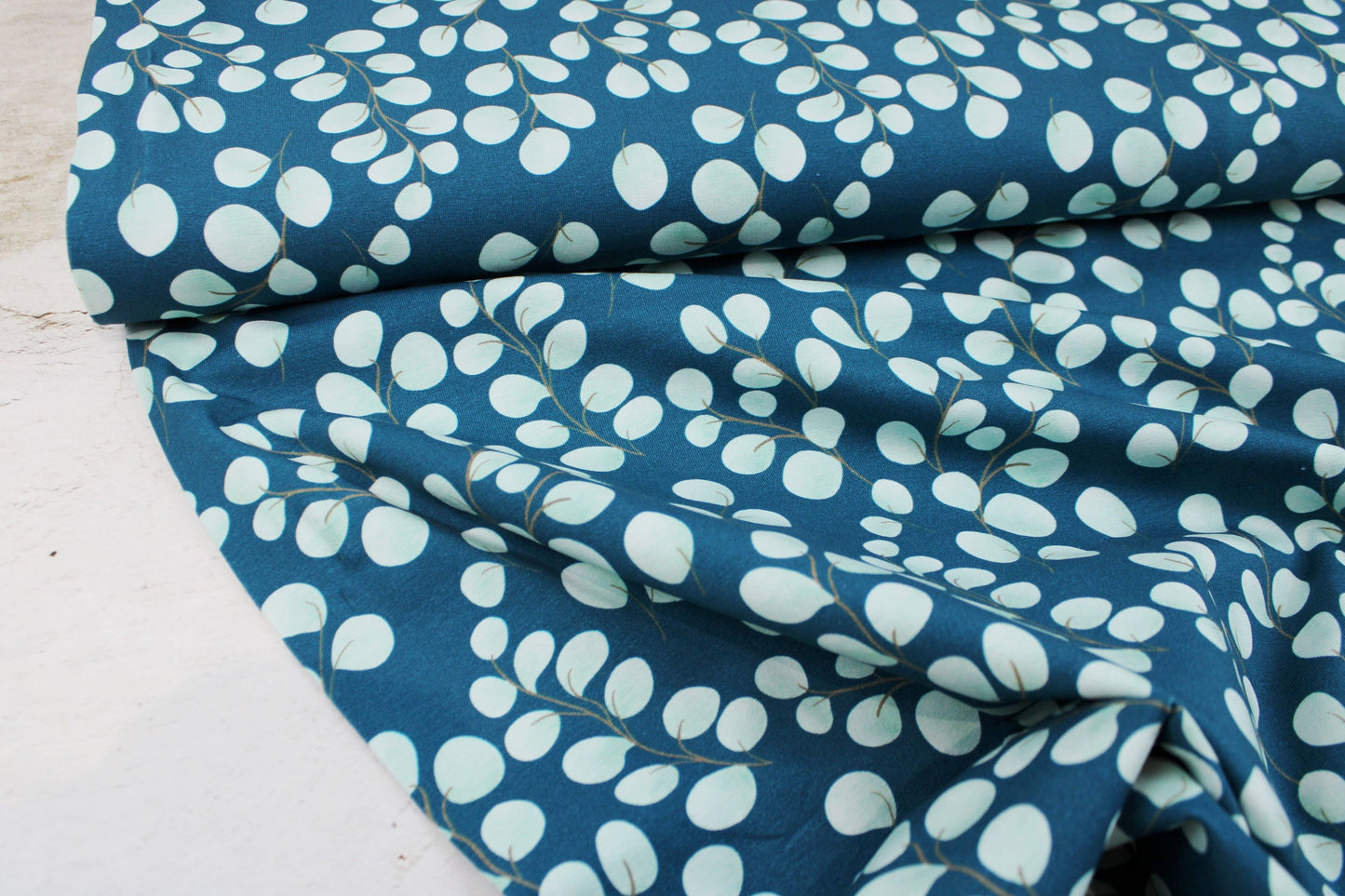 Baumwolljersey Fine Eukalyptus mit kleinen mintfarbenen Blättern auf petrol Grund Ballenansicht